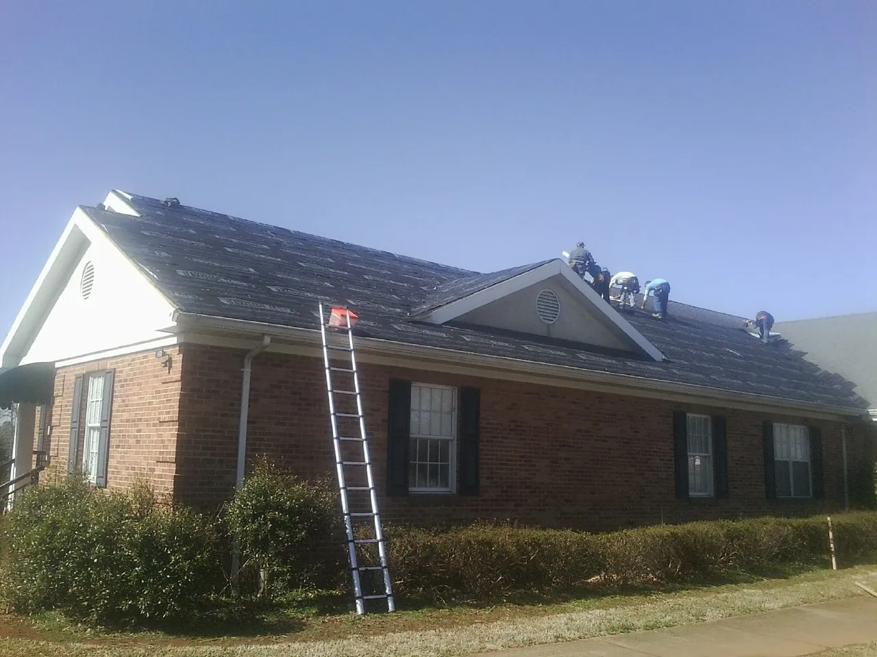 Crew tearing shingles off a brick ranch roof with a ladder set — Metal Roof Repair work by our crew in Wells Tannery