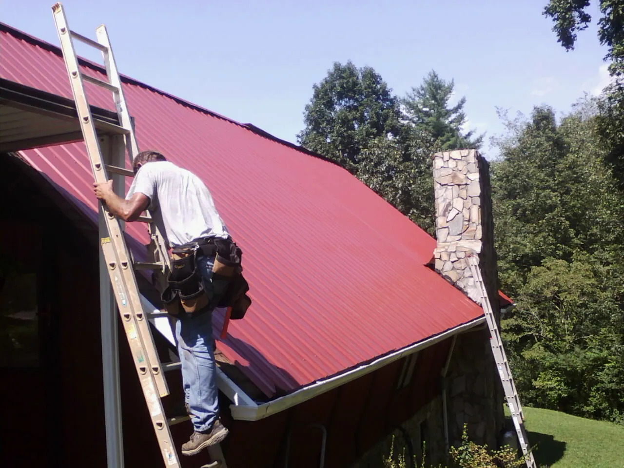 Roofer on a ladder beside a red metal roof and stone chimney — Soffit & Fascia Repair by Roofing Brothers in Wells Tannery