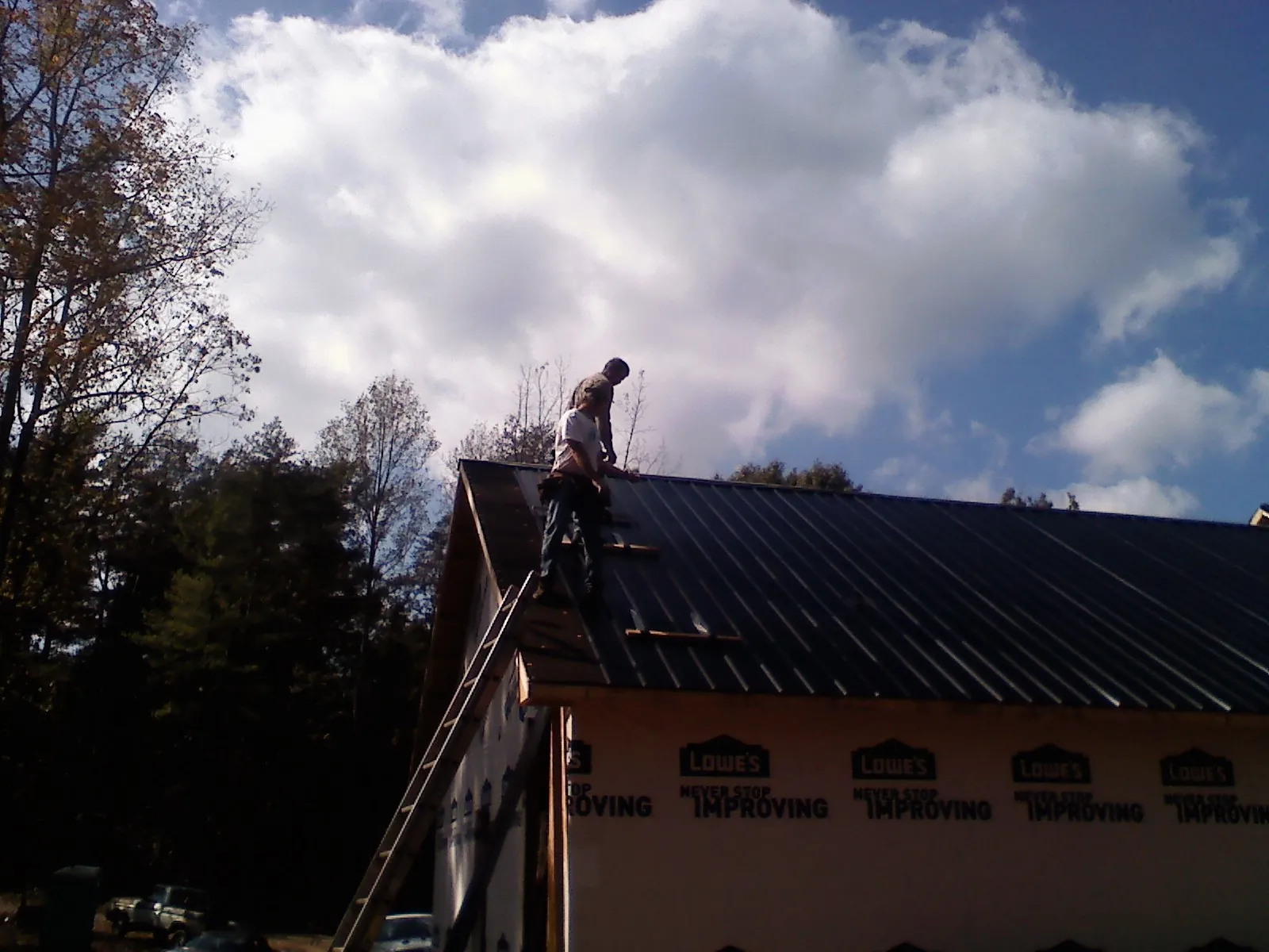 Roofer fastening dark metal panels high against a cloudy sky, from a Roof Inspection job in Wells Tannery